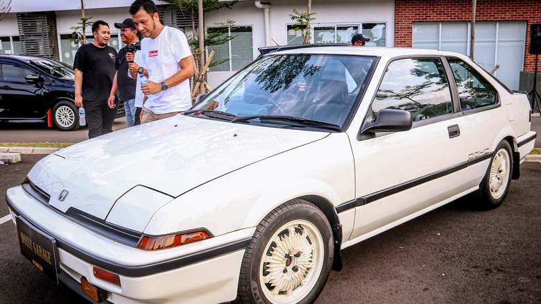 White first generation Integra coupe parked at a car meet, with custom wheels and several people gathered around in the background.