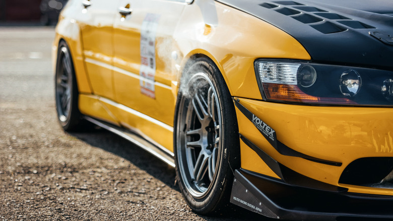 Close-up of a modified yellow race car with a black hood, showing smoke coming from the front wheel area.