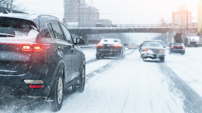 Cars driving on a snow-covered city road during a winter storm with brake lights illuminated and reduced visibility from falling snow.