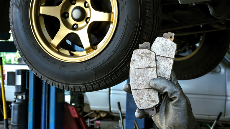 Mechanic holding two worn brake pads in front of a raised car with a gold-colored wheel in an auto repair shop.