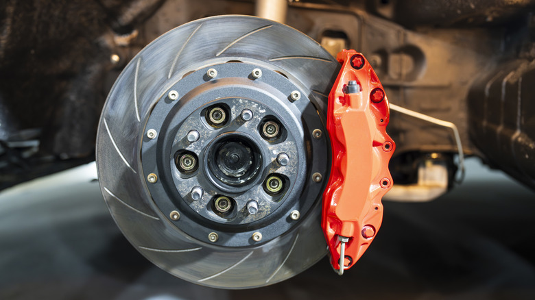 A high-performance slotted brake rotor with a bright red brake caliper mounted on a vehicle suspension system.