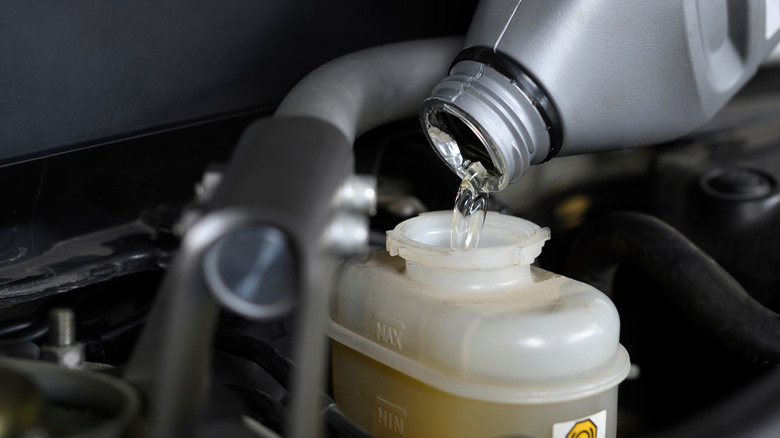 Brake fluid being poured from a gray plastic container into a car's brake fluid reservoir under the hood.