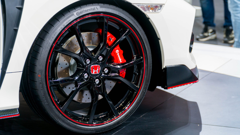 Close-up of a black and red Honda wheel with a visible Brembo brake caliper and drilled brake rotor on a white sports car.