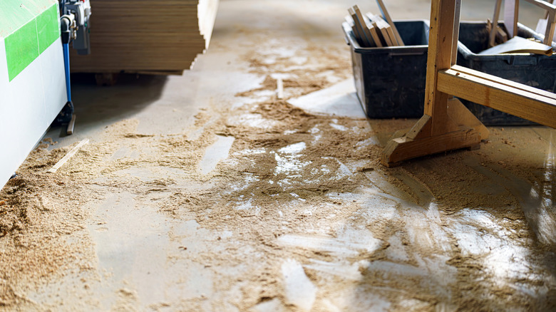 A shop floor with a lot of sawdust scattered around and bins, wood piles, and shop table legs in the shot.