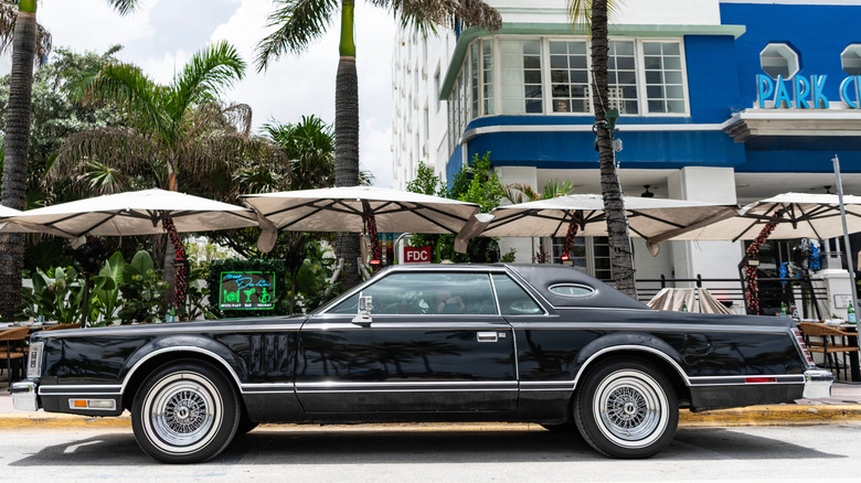 A black Mark V Lincoln Continental parked at the side of a roadside cafe, blue building in the background.