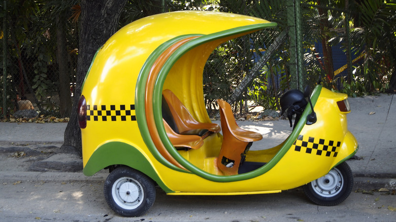 Bright yellow three-wheeled coconut-shaped taxi scooter parked on a tropical street in Havana, Cuba, with checkered detailing and open seating.