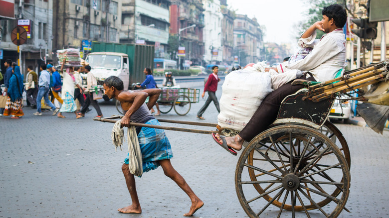 A man pulling a cart-rickshaw, with a passenger in the seat (both men unidentified) along a paved road.