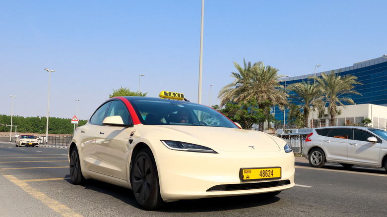Cream-colored Tesla Model 3 taxi with Dubai license plate and yellow rooftop taxi sign driving on a sunny urban road lined with palm trees and modern buildings.