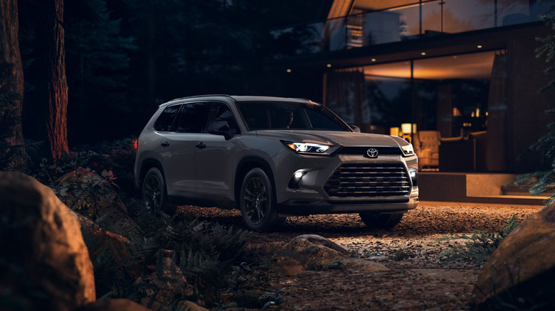 A grey 2025 Toyota Grand Highlander outside a modern log cabin, headlights on, front three quarters visible in the evening