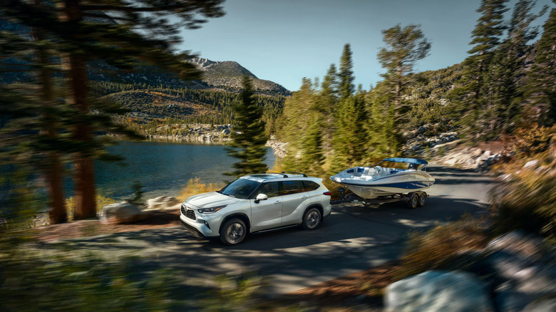 A satin white 2025 Toyota Highlander pulling a seadoo yacht along a forest road, with a lake visible in the background, in the afternoon.