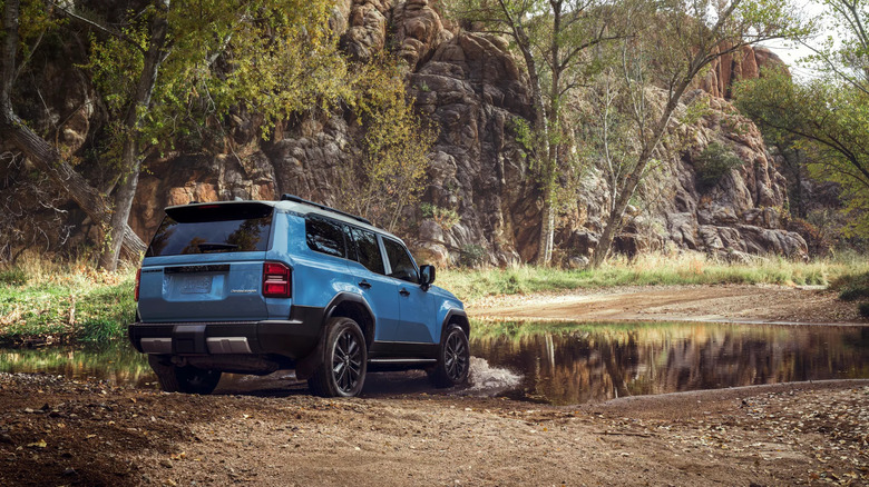 A blue 2025 Land Cruiser parked in a forest at the edge of a large puddle of water in the afternoon, read and right views visible of the car