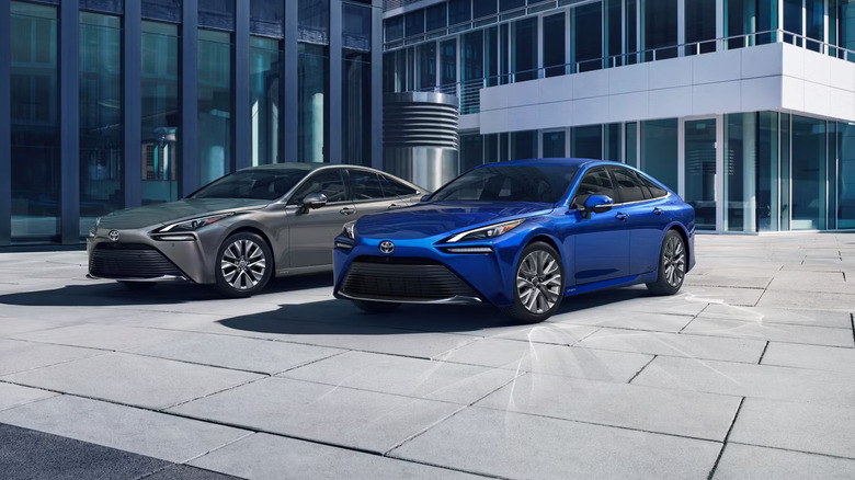 Two 2025 Toyota Mirai Fuel Cell EVs, one silver and one blue, parked against an office building, front and front-left views of the cars visible.
