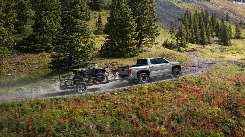 A silver-grey Toyota Tacoma pulling a trailer with a quad-bike ATV in it up a hilly gravel road, with green fields and hills visible in the background