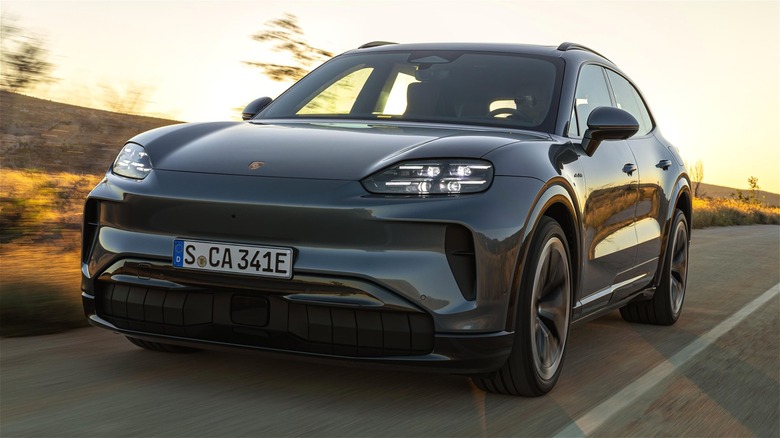 A dark gray 2026 Porsche Cayenne EV cruising along a winding rural road at golden hour against the hillside backdrop.