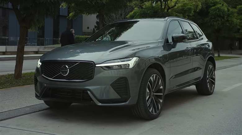 A gray 2027 Volvo EX60 is parked on a tree-lined city street beside modern office buildings.