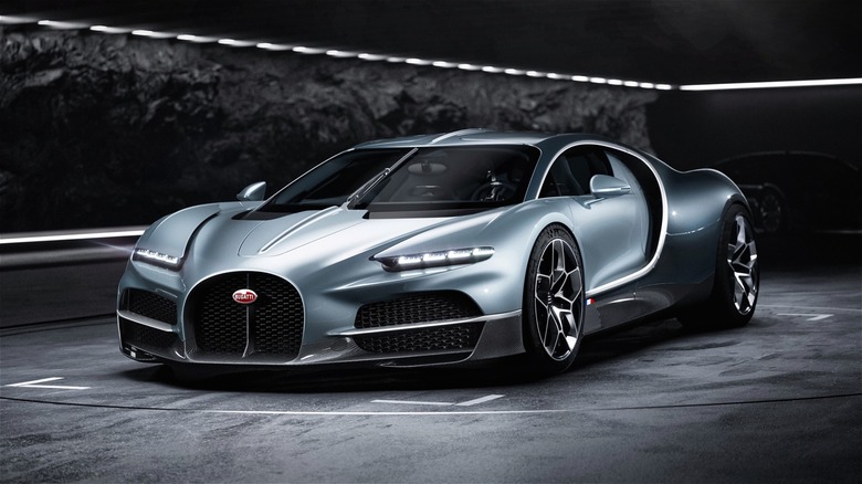 A sleek silver 2026 Bugatti Tourbillon hypercar sits dramatically in a tunnel-like studio under soft overhead lighting in a dark.