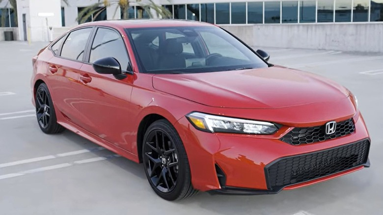 A 2025 red Honda Civic Hybrid sedan parked in a modern rooftop lot with palm trees and glass buildings in the background.