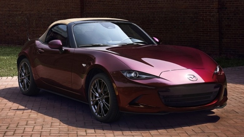 A 2025 deep red Mazda MX-5 Miata convertible parked in front of a brick building under soft light.