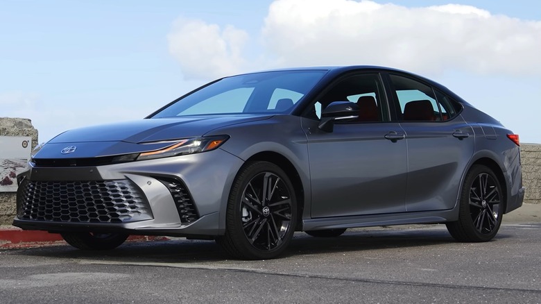 A 2025 gray Toyota Camry parked near a stone wall by the coast under a bright blue sky.