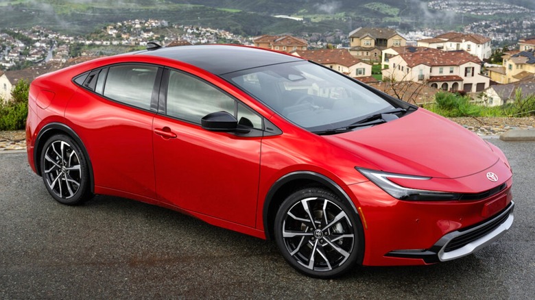 A 2025 red Toyota Prius parked on a scenic overlook with rolling hills, misty skies, and a suburban neighborhood in the background.