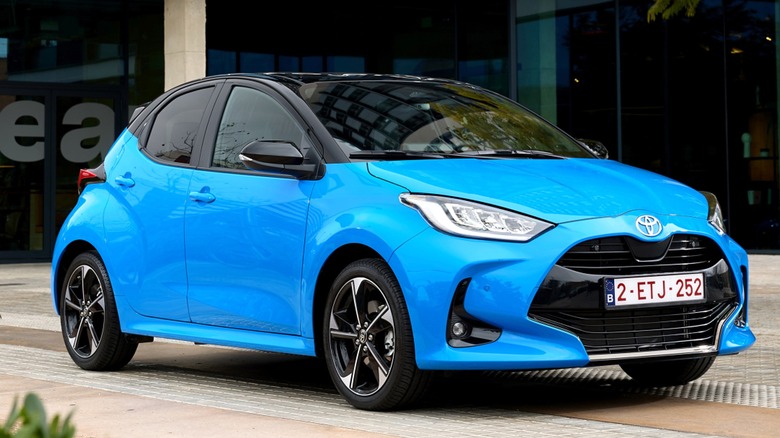 A 2024 bright blue Toyota Yaris parked in front of a modern glass building surrounded by greenery.