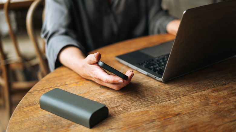 Person seated at a wooden table on a laptop holding a smartphone