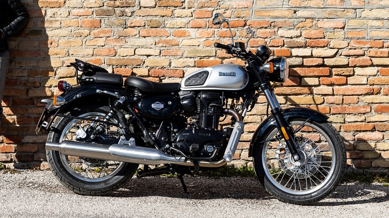 A Benelli Imperiale 400 motorcycle in classic silver and black, parked beside a brick wall with a rider in a black leather jacket standing casually nearby.