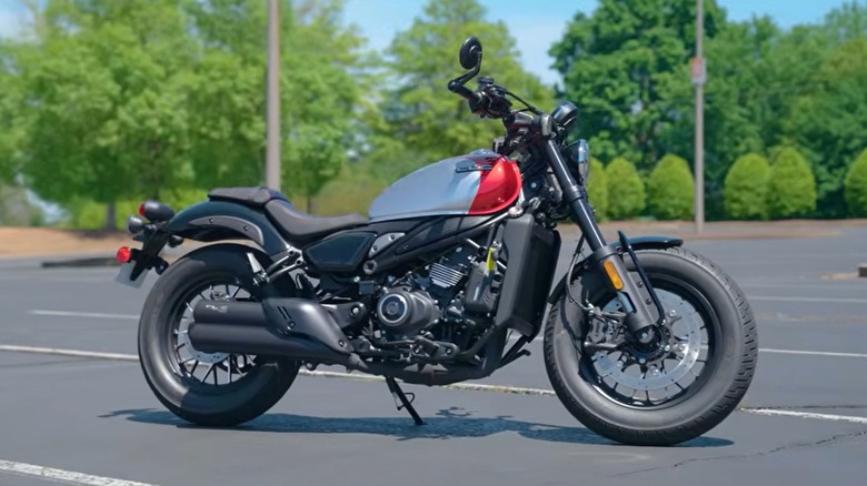 A CFMoto 450 CL-C motorcycle in a silver and red color scheme, parked in an empty paved lot with green trees in the background.