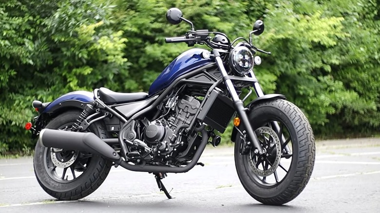 A Honda Rebel 300 motorcycle in blue, parked on pavement with greenery in the background.