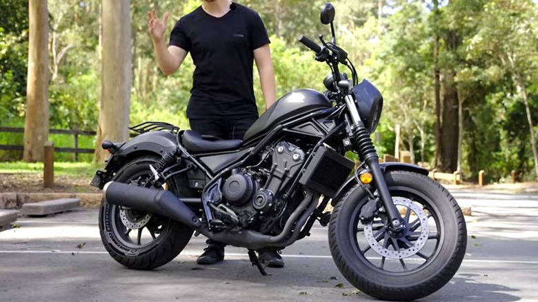 A black Honda Rebel motorcycle parked on a quiet road, with a rider dressed in black standing beside it with a backdrop of tall trees and greenery.