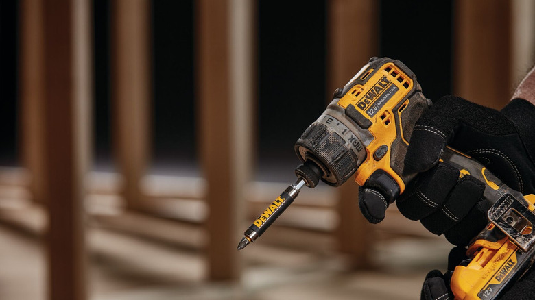 A person wearing black work gloves holds a compact yellow-and-black DeWalt brushless screwdriver.