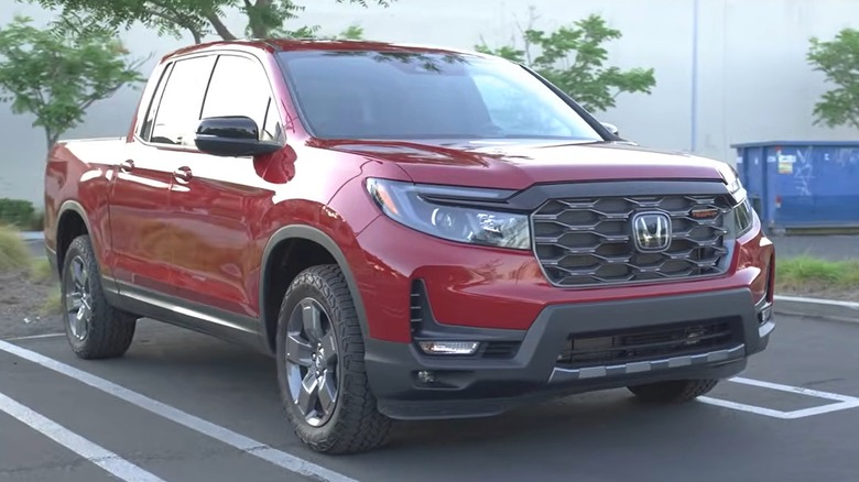 A red Honda Ridgeline pickup truck parked in a lot, with trees, a large building, and a blue Republic Services dumpster visible in the background.