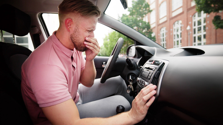 A man in the driver's seat of a car, holding his nose after catching a whiff of a bad smell.