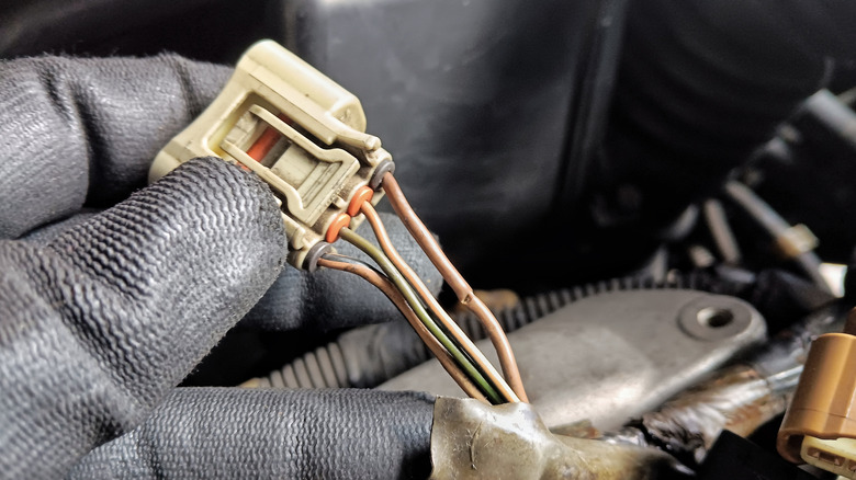 Close-up of a gloved hand holding a car wiring connector with exposed brown and green wires inside an engine bay.