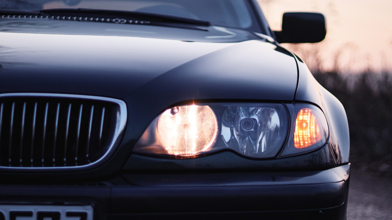 A close-up of the front of a dark-colored car with its headlights and turn signal on at dusk.