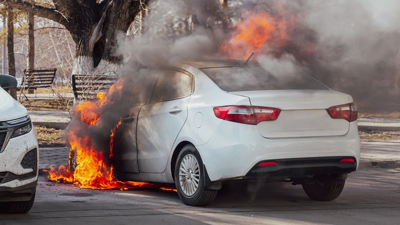 A white car parked outdoors with flames and thick smoke coming from the engine area.