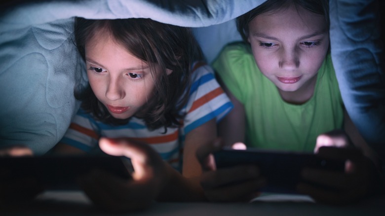 two kids playing on handheld gaming consoles under a blanket.
