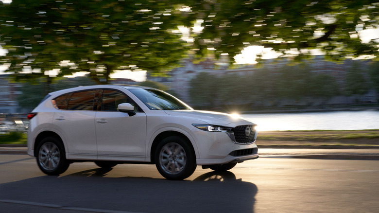 A white 2022 Mazda CX-5 driving along a tree-lined road near the water.