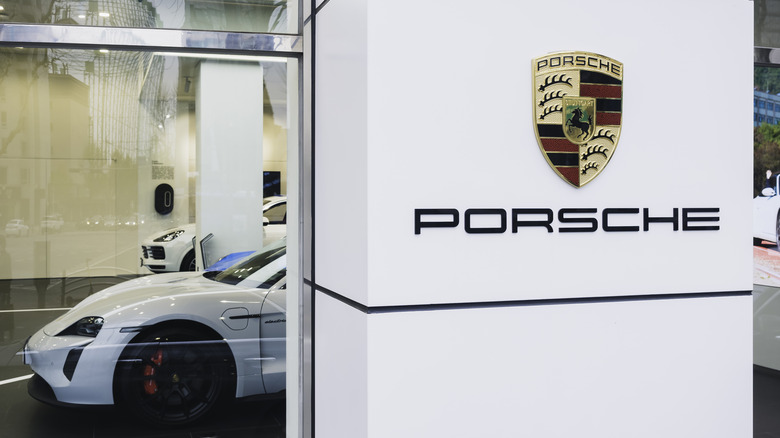 The Porsche logo and shield on a white pillar outside a Porsche dealership, car visible in the back.