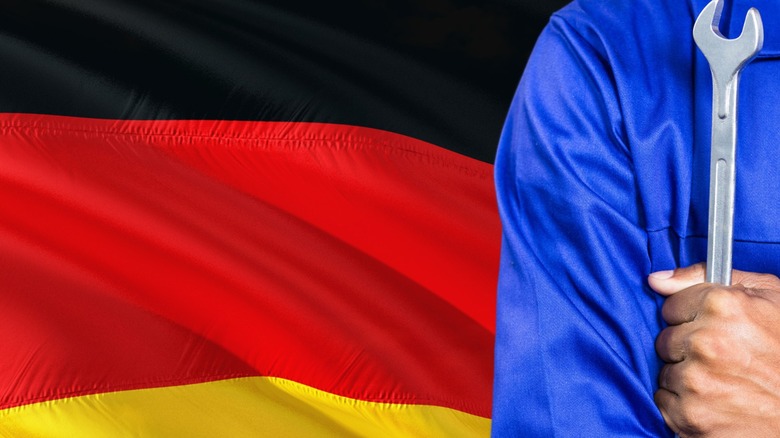 Side-shot of a man holding a spanner in blue overalls standing behind a flapping German flag