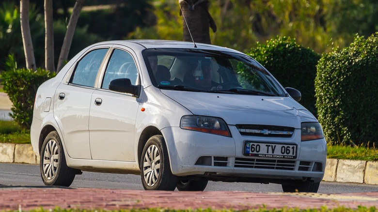 A white Chevrolet Aveo parked on the road with hedges and trees in background