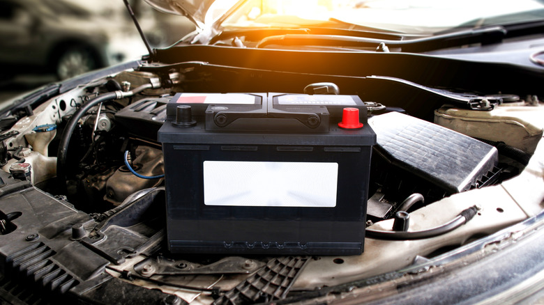 A black car battery sitting on a car's engne bay with sun shining behind it.