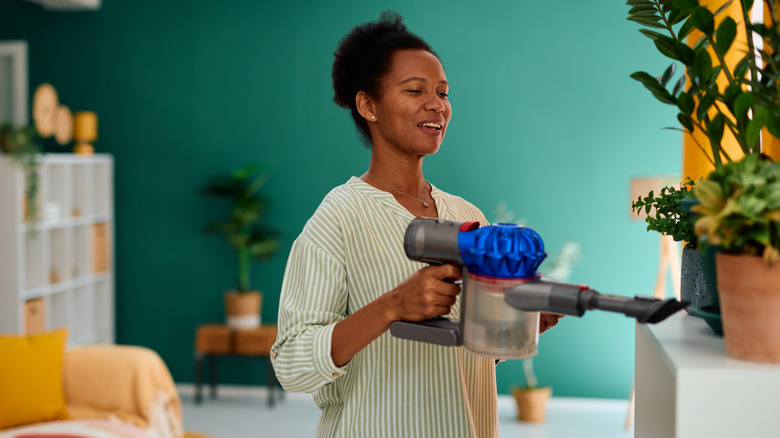 A smiling African American woman uses a handheld vacuum to clean a white shelf with houseplants on it