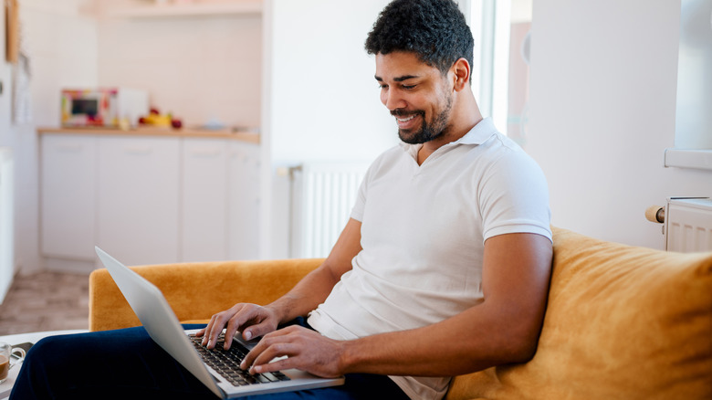 A young man smiling while working on a laptop which is on his lap