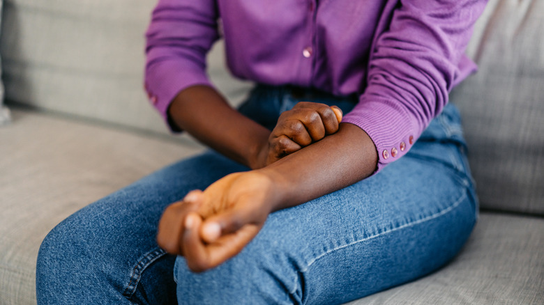 Person in a purple shirt and blue jeans scratching at their forearm.