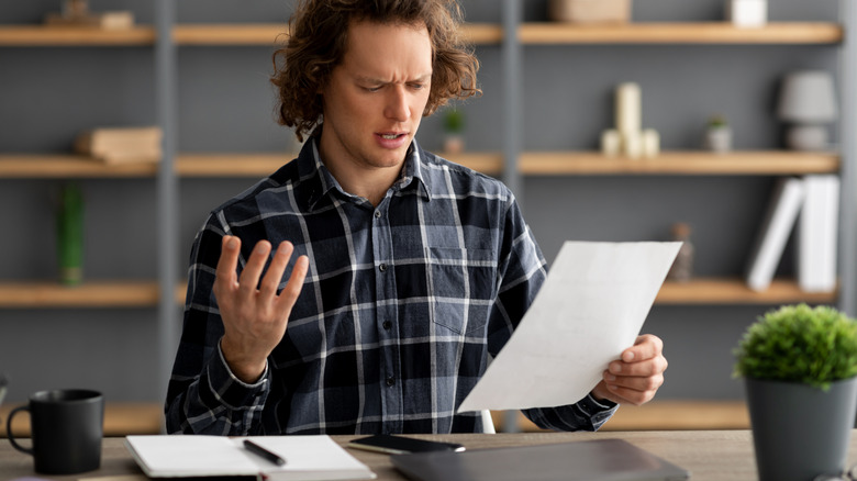 Man sitting at a laptop looking at a piece of paper and appearing confused.