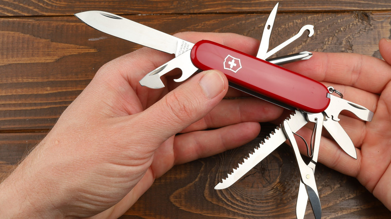 Hands holding a Swiss Army Knife with numerous tools open, including the blade, can opener, hook, scissors and saw.