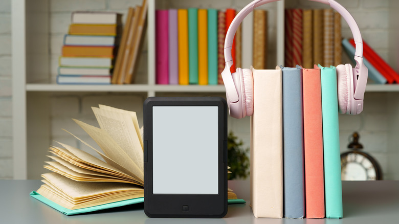 An e-reader next to an open book and four books held upright by a pair of pink headphones