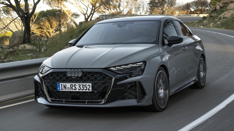 A gray Audi RS3 sedan on a two-lane road, surrounded by trees and rocky hills.