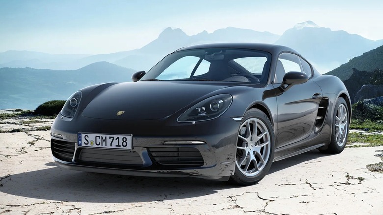 A sleek dark gray Porsche 718 Cayman posed on a rocky coastal landscape with mountains behind it.